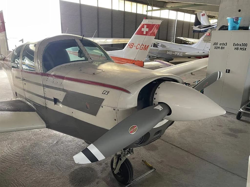 A front view of the white and red single-engine aircraft, HB-DET, showing its propeller and nose inside a hangar.