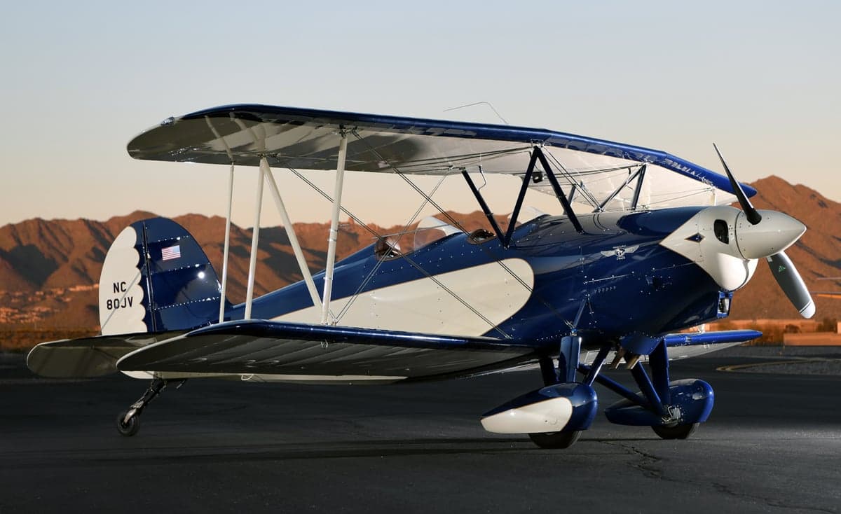 A blue and white biplane, N80JV, is parked on the tarmac with mountains in the background.