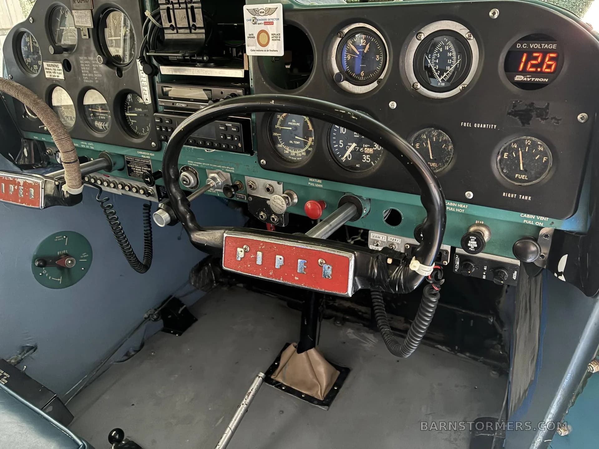 The cockpit of a Piper aircraft showing the instrument panel with a black control yoke featuring a red "PIPER" emblem.