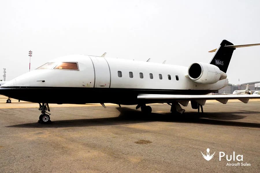 A side view of a white and black Bombardier Challenger 604 business jet (N64JS) on a tarmac.