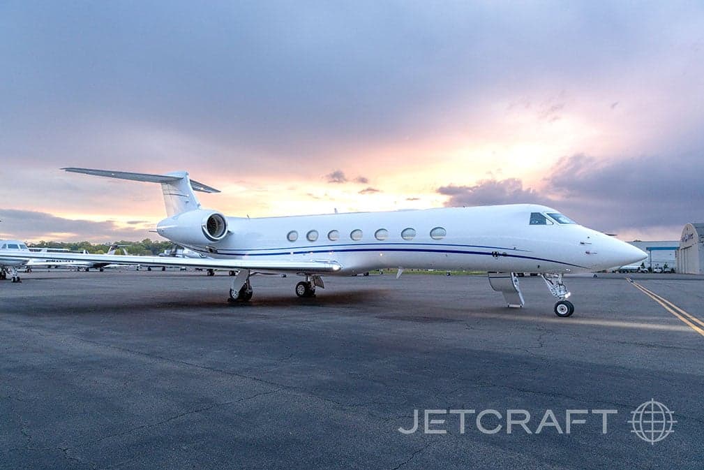 A white Gulfstream G550 business jet with blue stripes parked on a tarmac under a cloudy sky.
