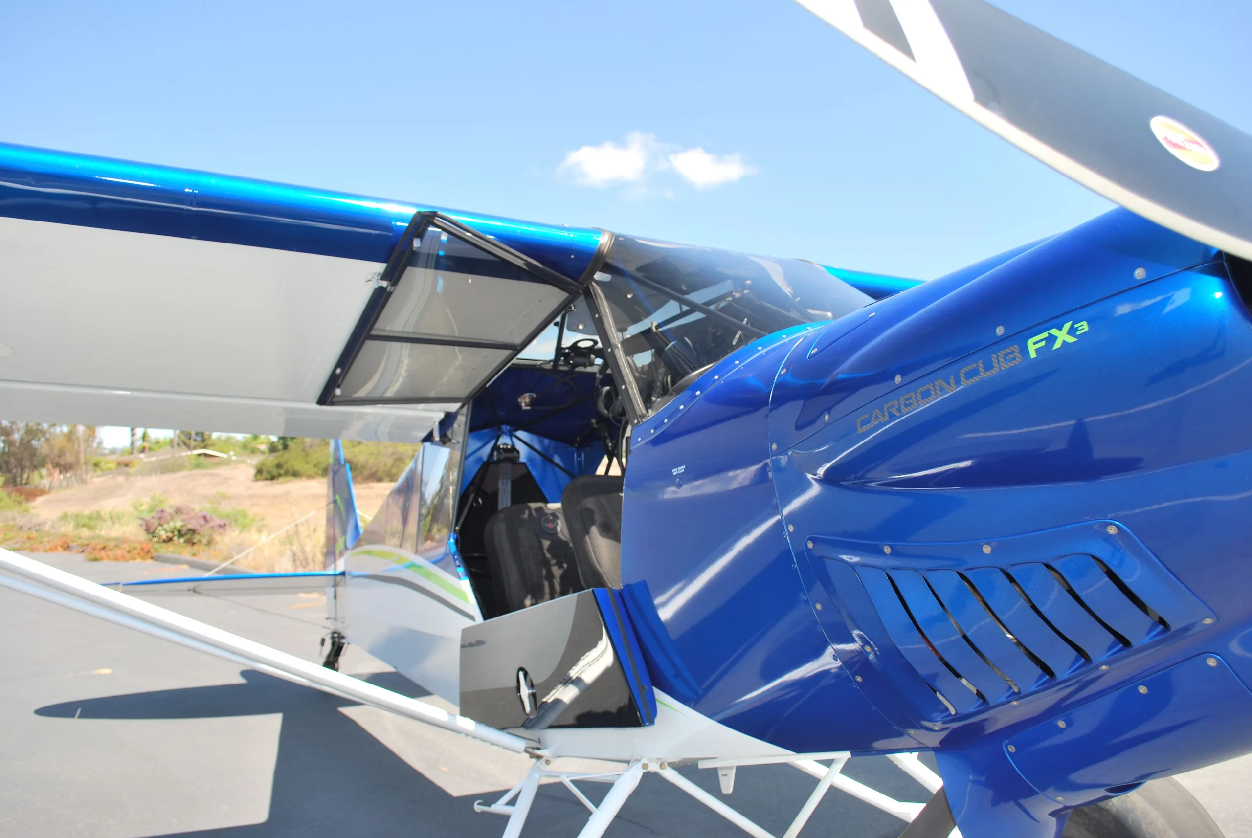 A side view of a blue and white Carbon Cub FX3 aircraft with its door open, parked on an asphalt ramp.