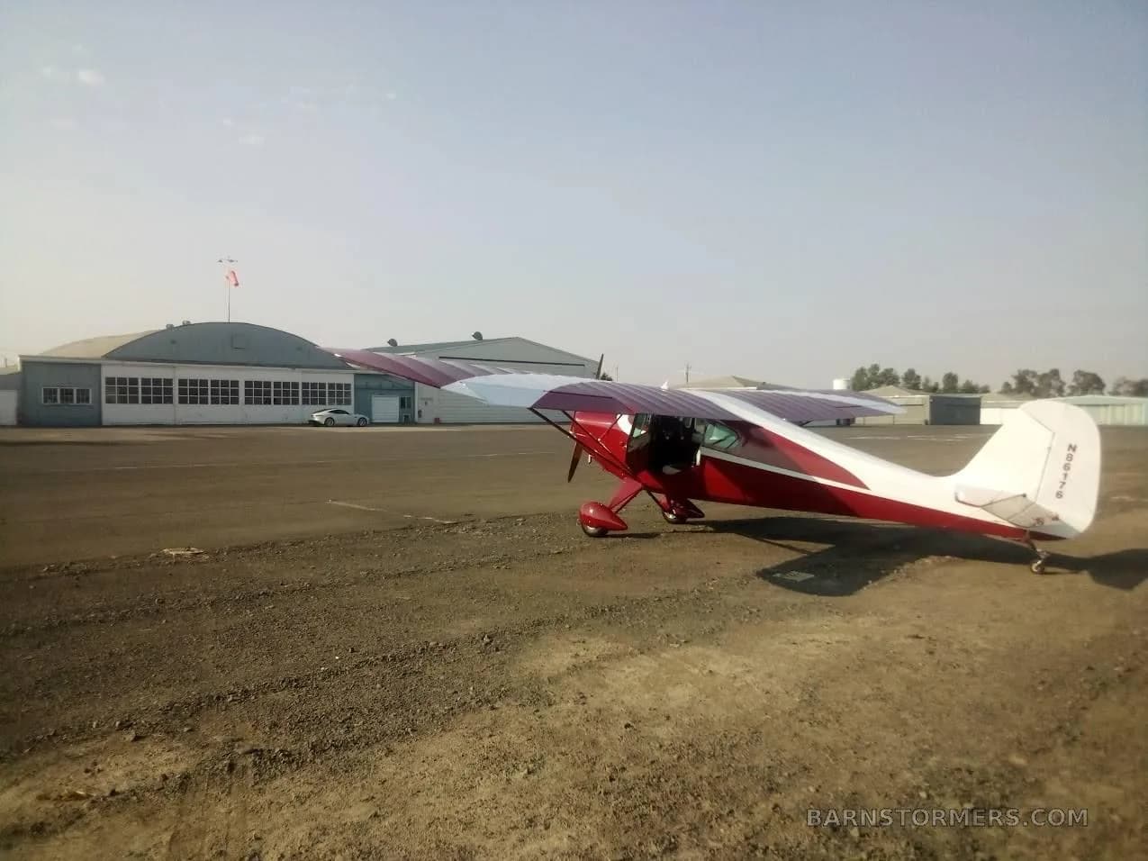 A red and white Aeronca Chief N86176 parked on a dirt and asphalt airfield with hangars in the background.
