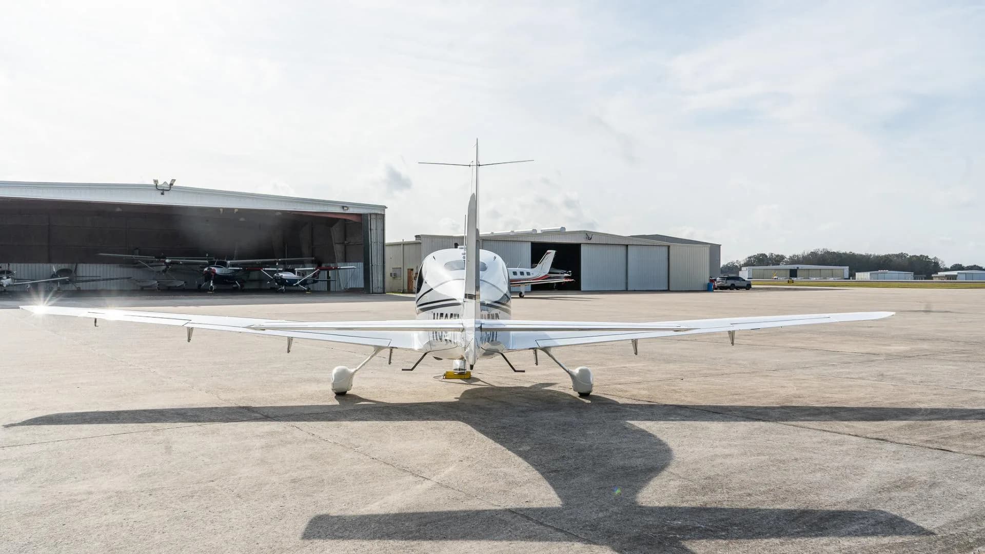 Rear view of a white Cirrus SR22 aircraft parked on an airfield with hangars in the background.