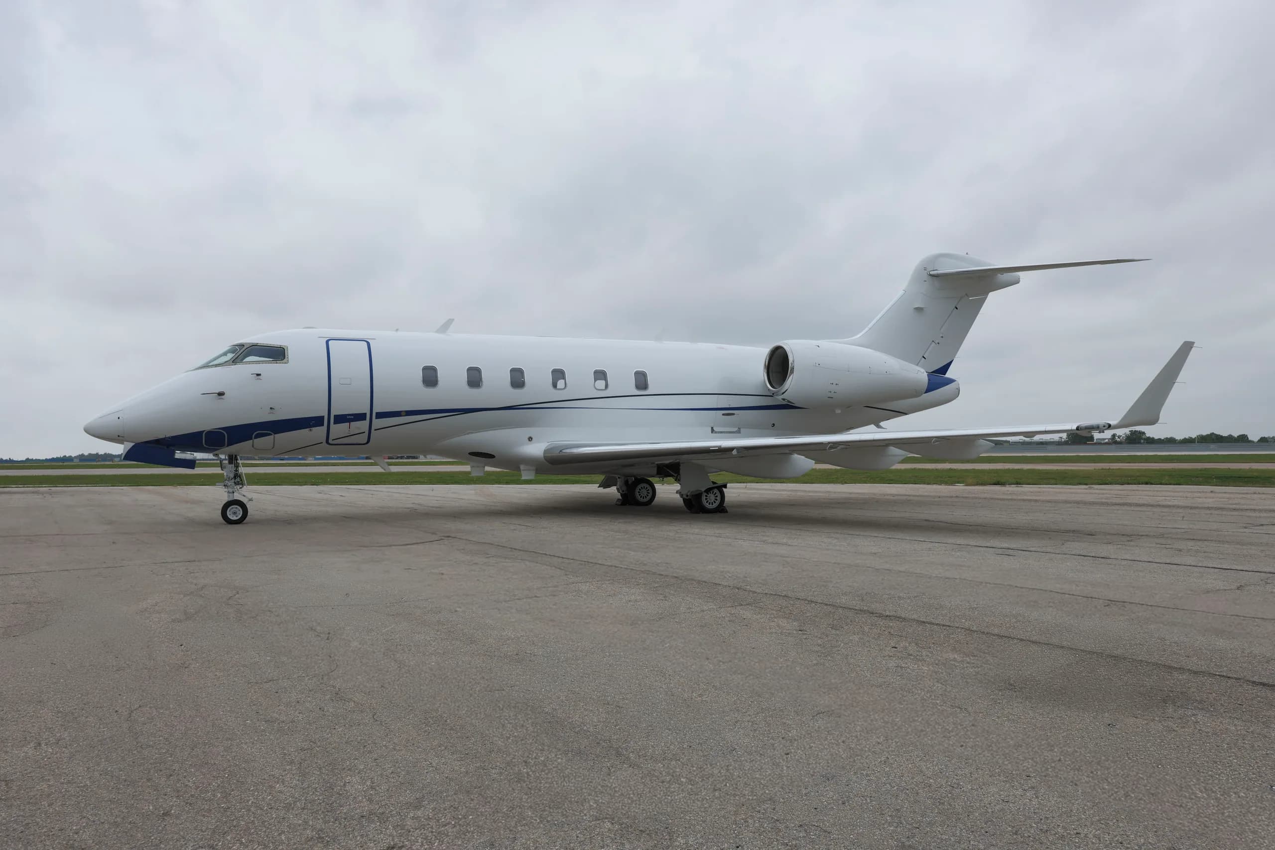 A white Bombardier Challenger business jet with blue accent stripes parked on an airport tarmac under a cloudy sky.