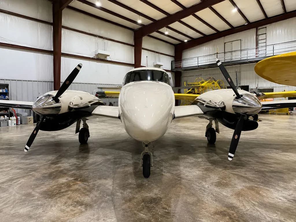 A head-on view of a white and black twin-engine propeller aircraft parked in a hangar.