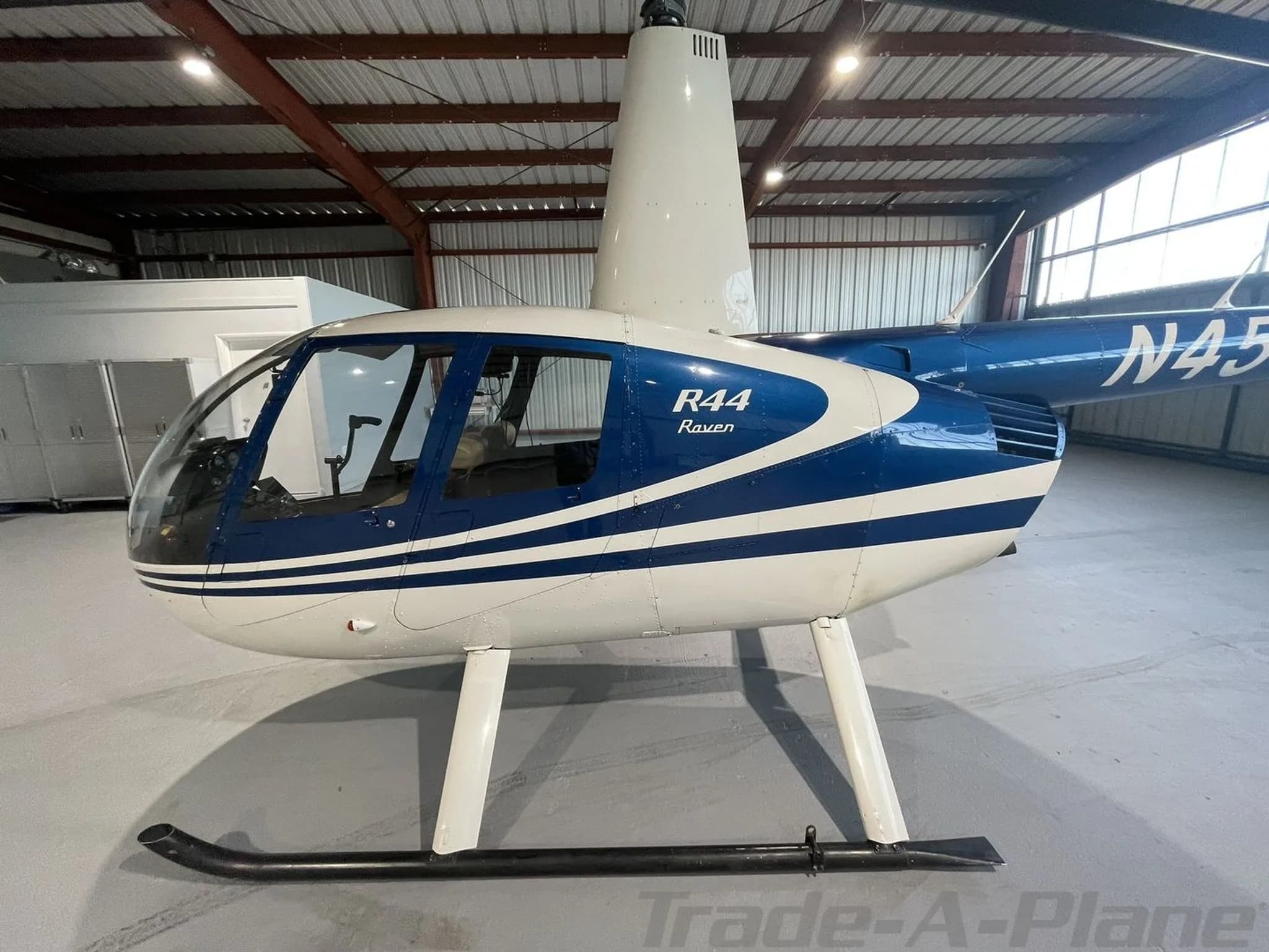 A close-up side view of the blue and white Robinson R44 Raven helicopter's cockpit and fuselage in a hangar.