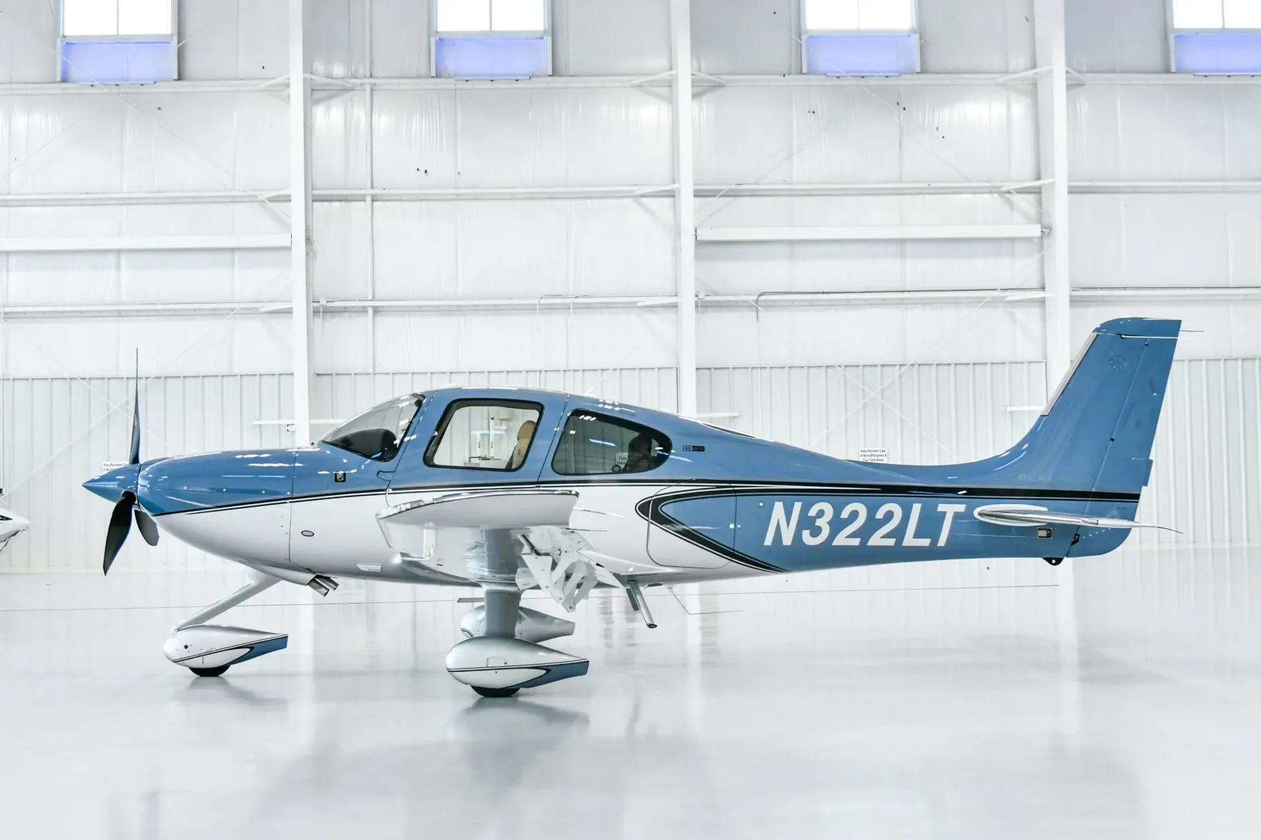 A blue and white Cirrus SR22 aircraft with registration N322LT parked in a brightly lit hangar, viewed from the side.