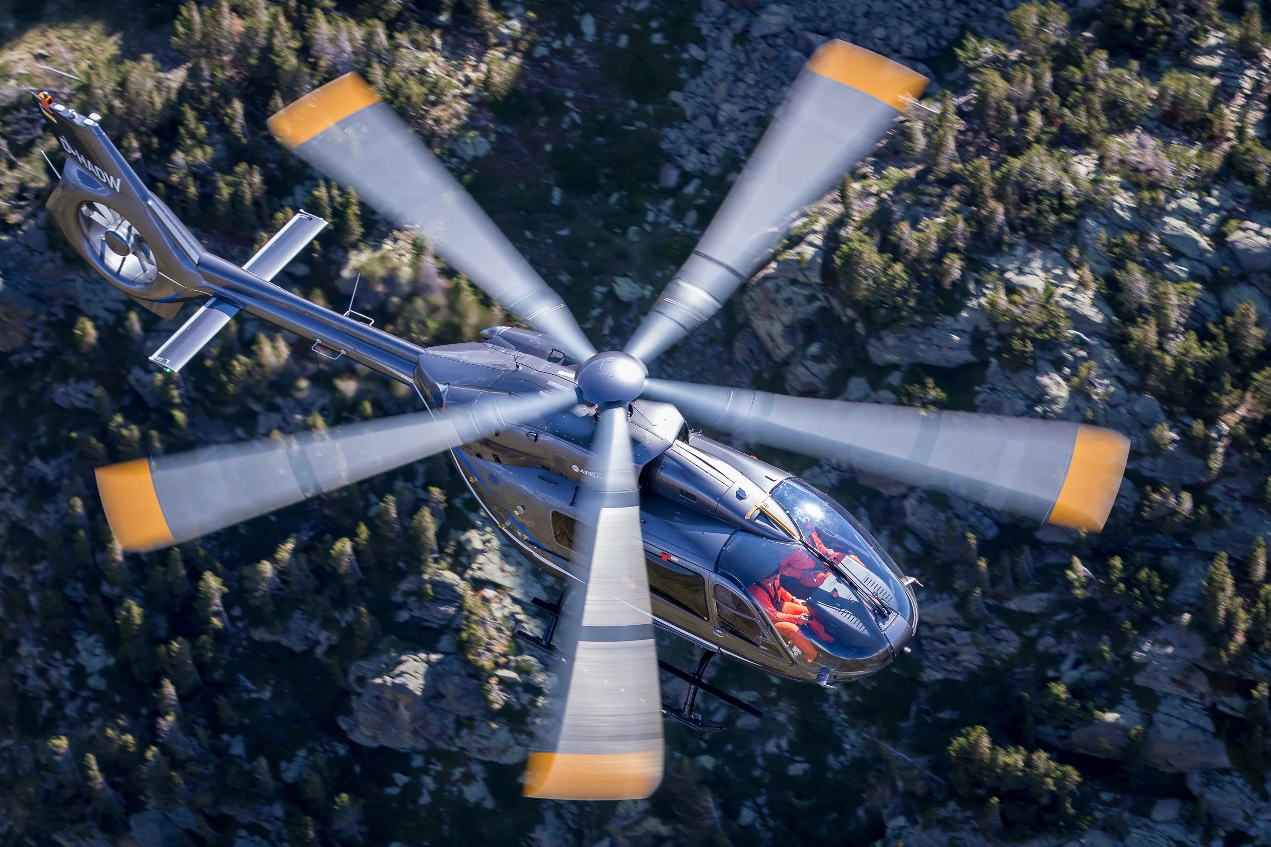 A dark gray helicopter with orange accents flying over a forest.