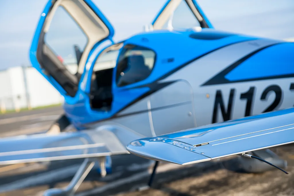 A close-up, blurred view of the blue and silver Cirrus SR22T GTS Carbon aircraft (N127DF) with its doors open, focusing on the wing.