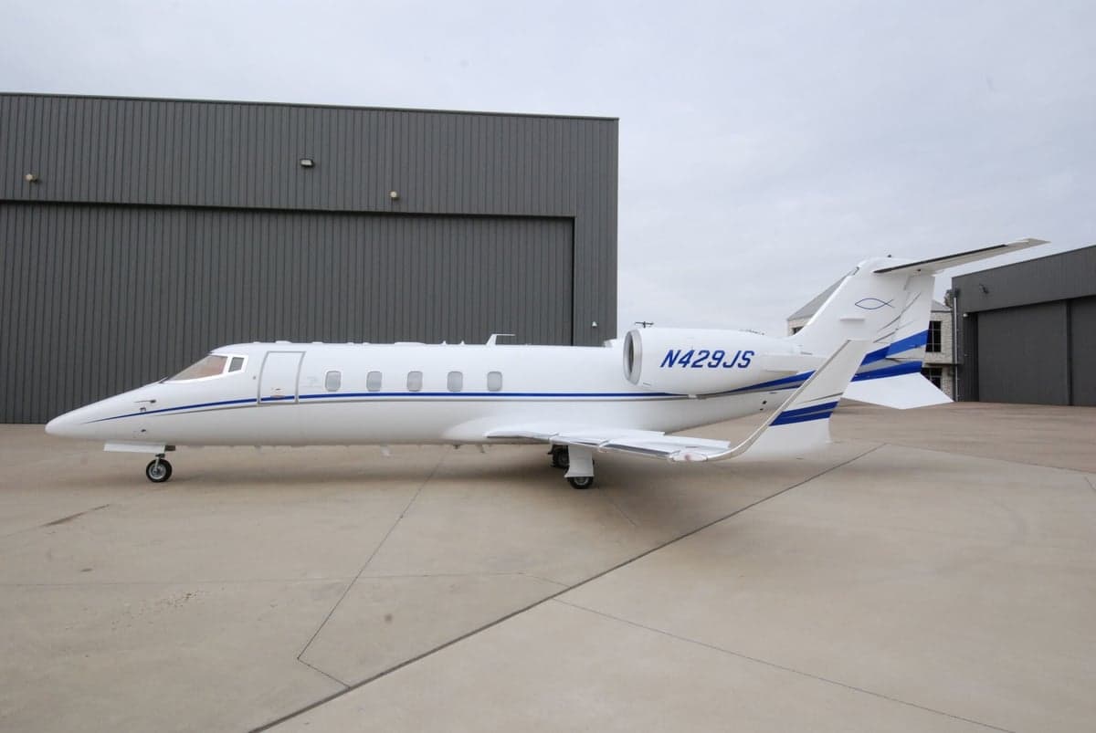A white business jet with blue stripes and registration N429JS parked on a tarmac in front of dark hangars.