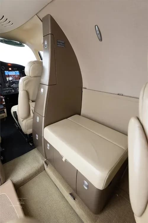 A view inside the Cessna Citation M2 cabin showing a beige bench seat and storage compartments, with the cockpit visible in the background.