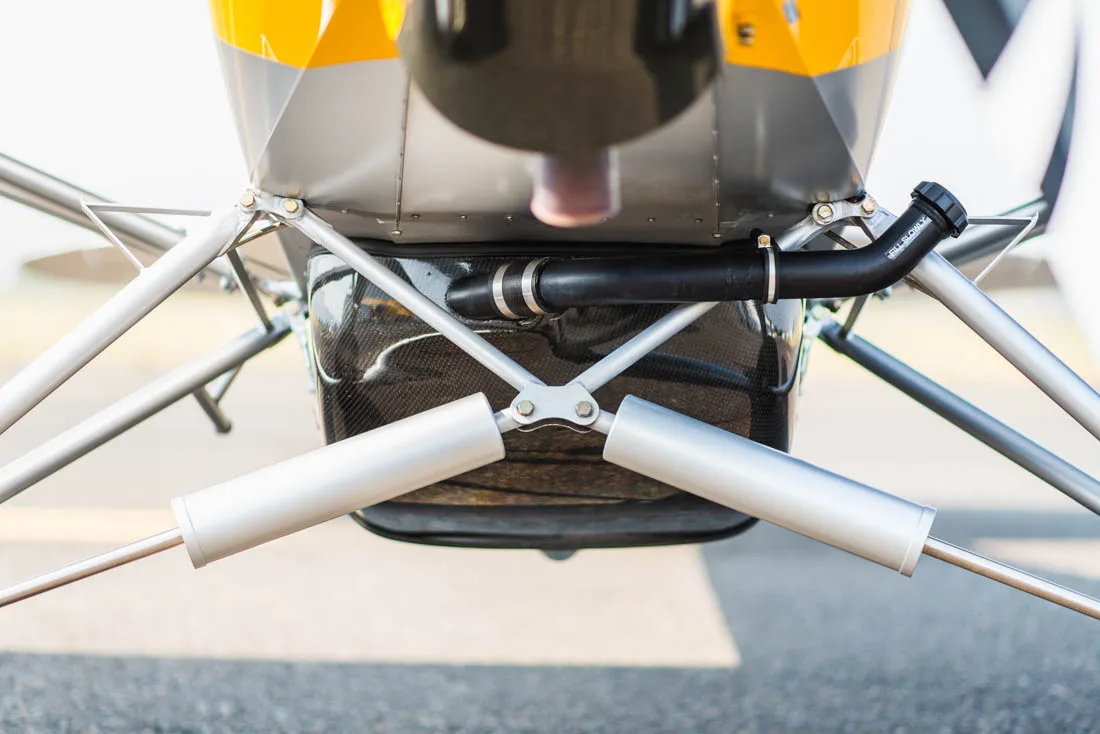 A close-up view of the underside of a Carbon Cub fuselage shows the exhaust pipe and landing gear struts.