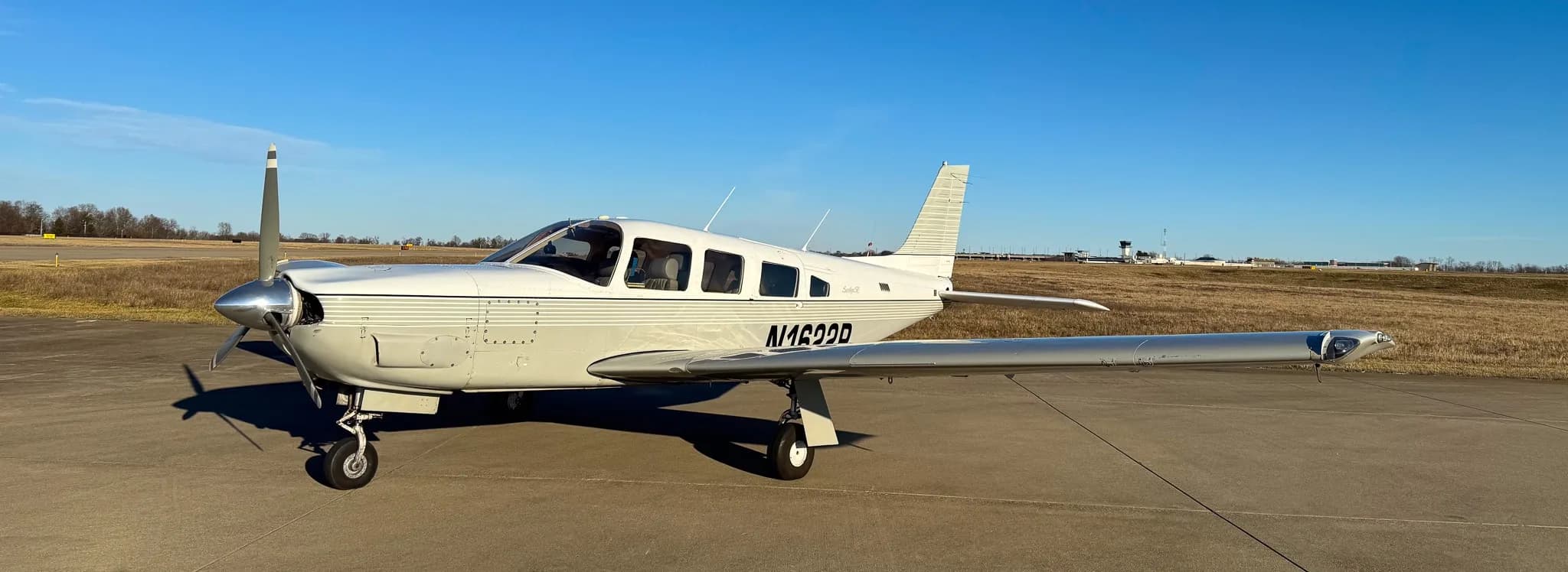 A white Piper Saratoga SP (N1622P) with grey stripes parked on an airfield under a clear blue sky.