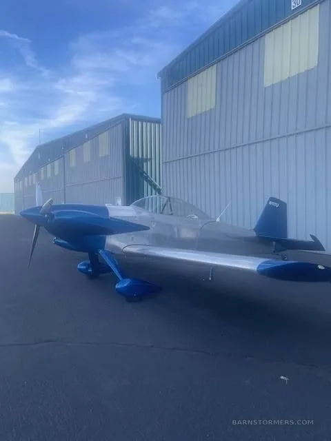A blue and silver single-engine aircraft with a propeller, viewed from the front-right, parked next to hangars under a blue sky.