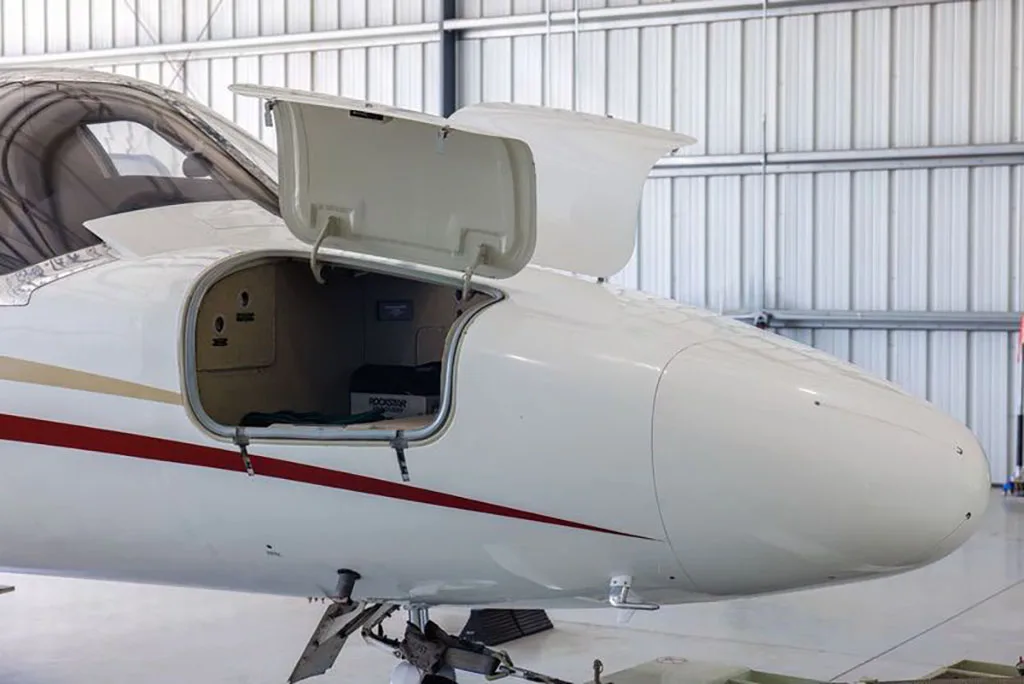 The nose of a white business jet with red and gold stripes, showing an open front baggage compartment inside a hangar.