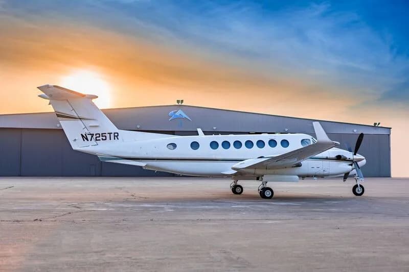 A white Beechcraft King Air 350i with tail number N725TR is parked next to a hangar at sunset.