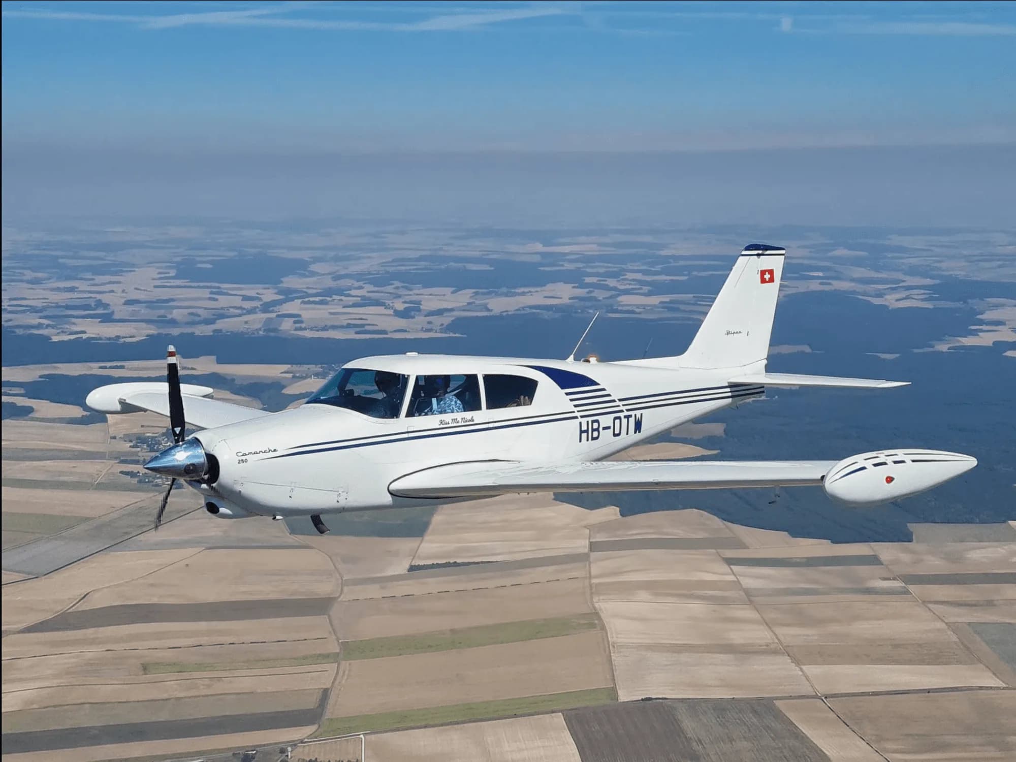 A Piper Comanche 250 in flight over a rural landscape.