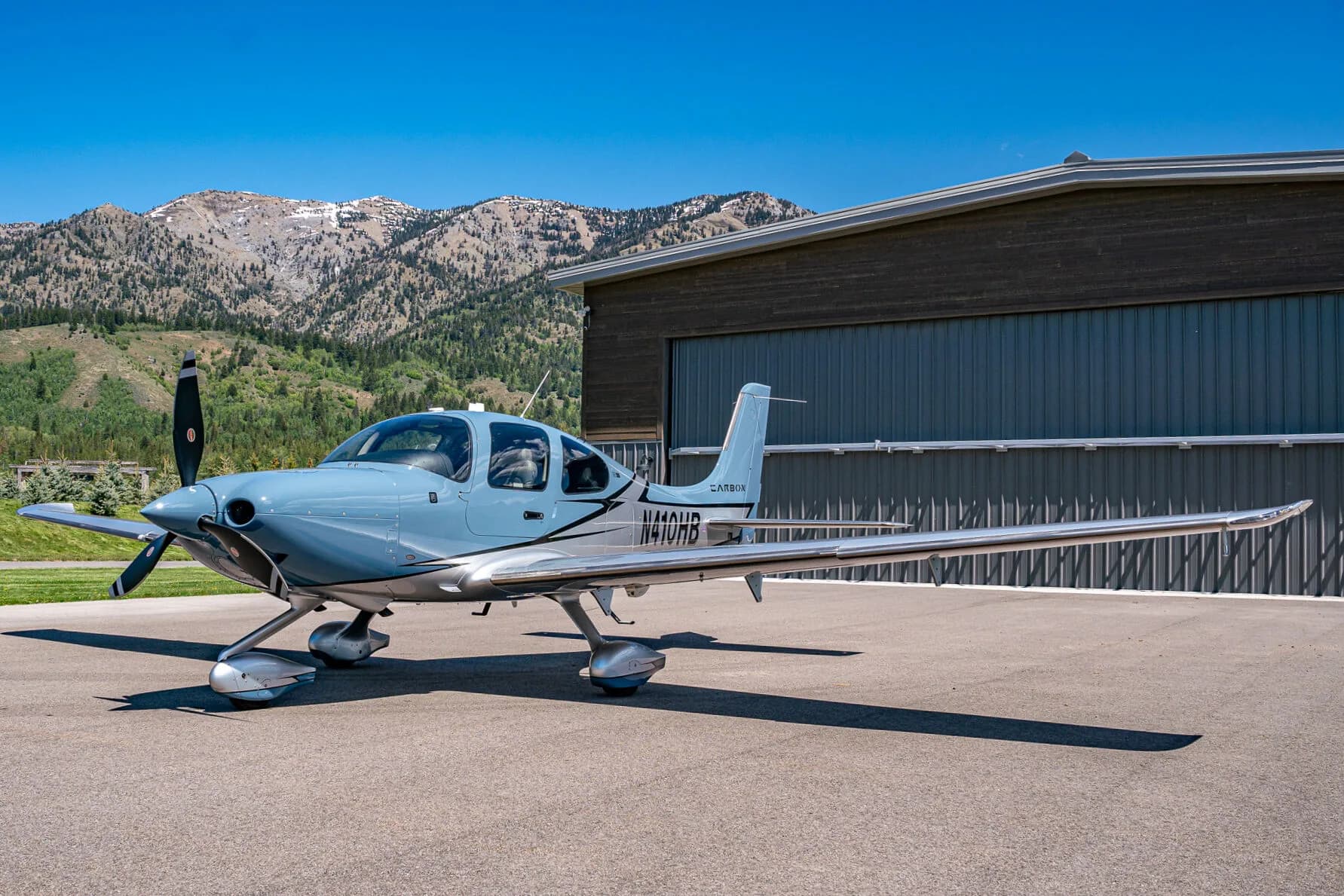 A light blue and silver Cirrus SR22T aircraft (N410HB) parked next to a hangar with mountains in the background.