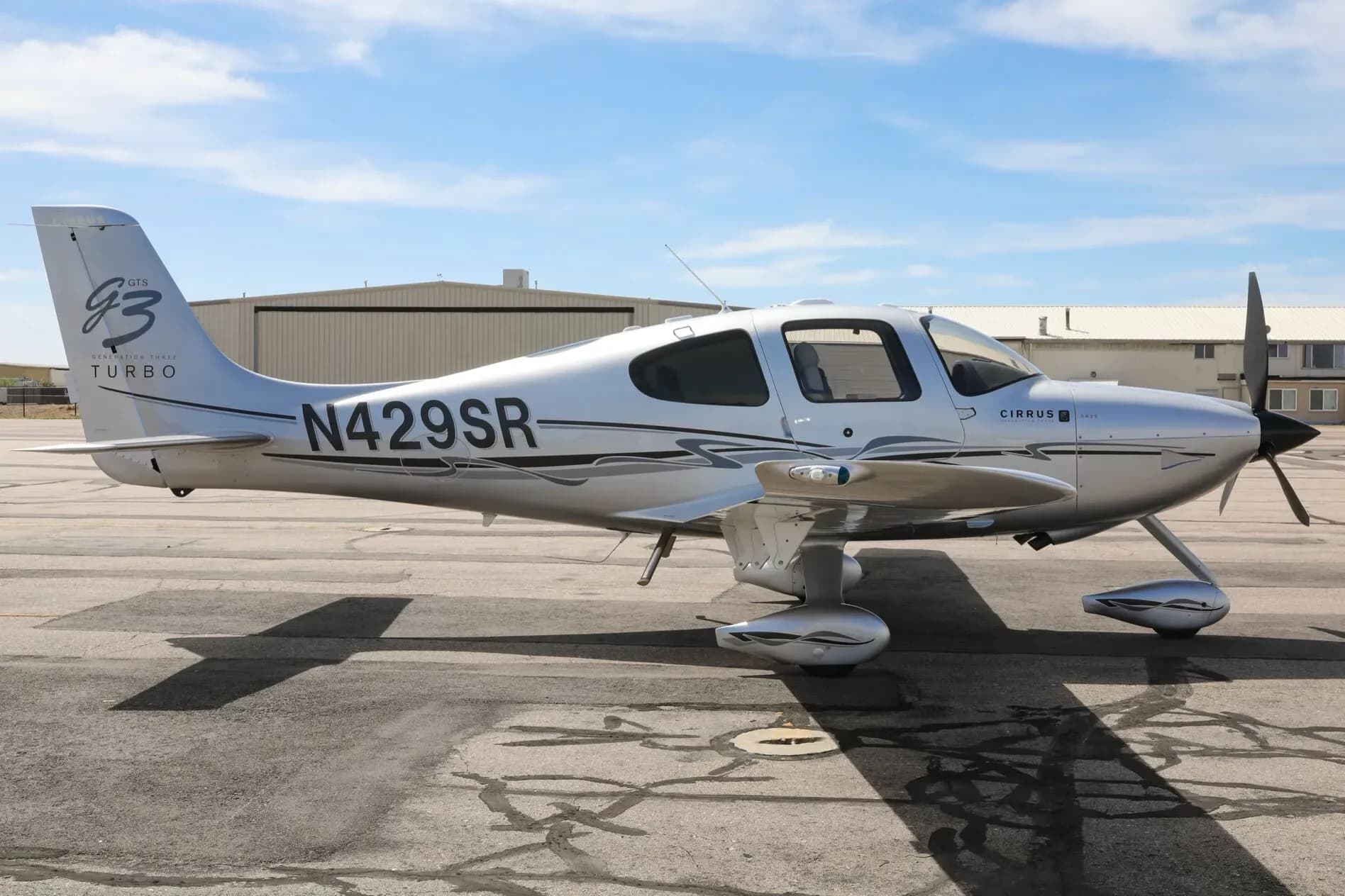A silver Cirrus SR22T aircraft with black and grey accents, registration N429SR, parked on an asphalt ramp with hangars in the background.