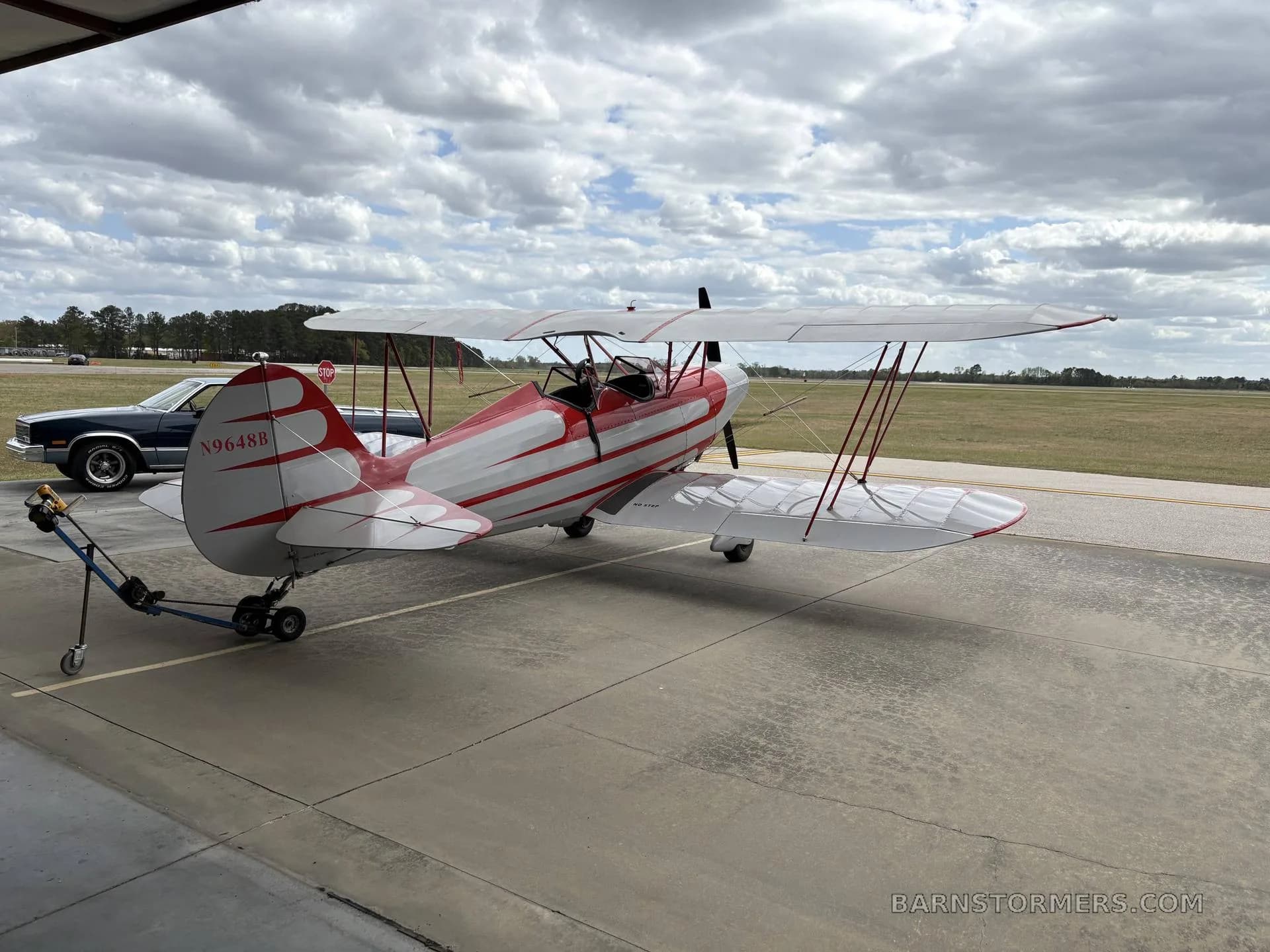 A red and white biplane with tail number N9648B is parked on an airfield tarmac under a cloudy sky.