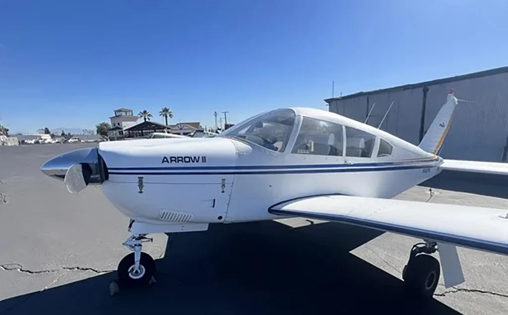 Front-left view of a white Piper Arrow II aircraft with blue stripes parked on an asphalt tarmac.