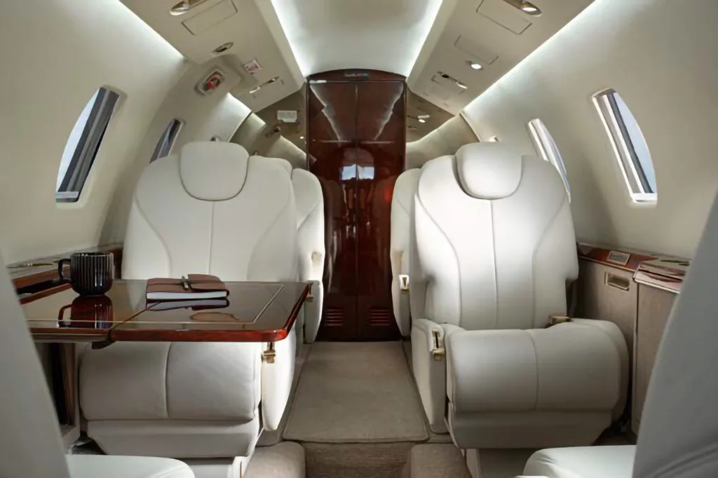 The luxurious interior of a private jet cabin with cream leather seats and a polished wooden table.