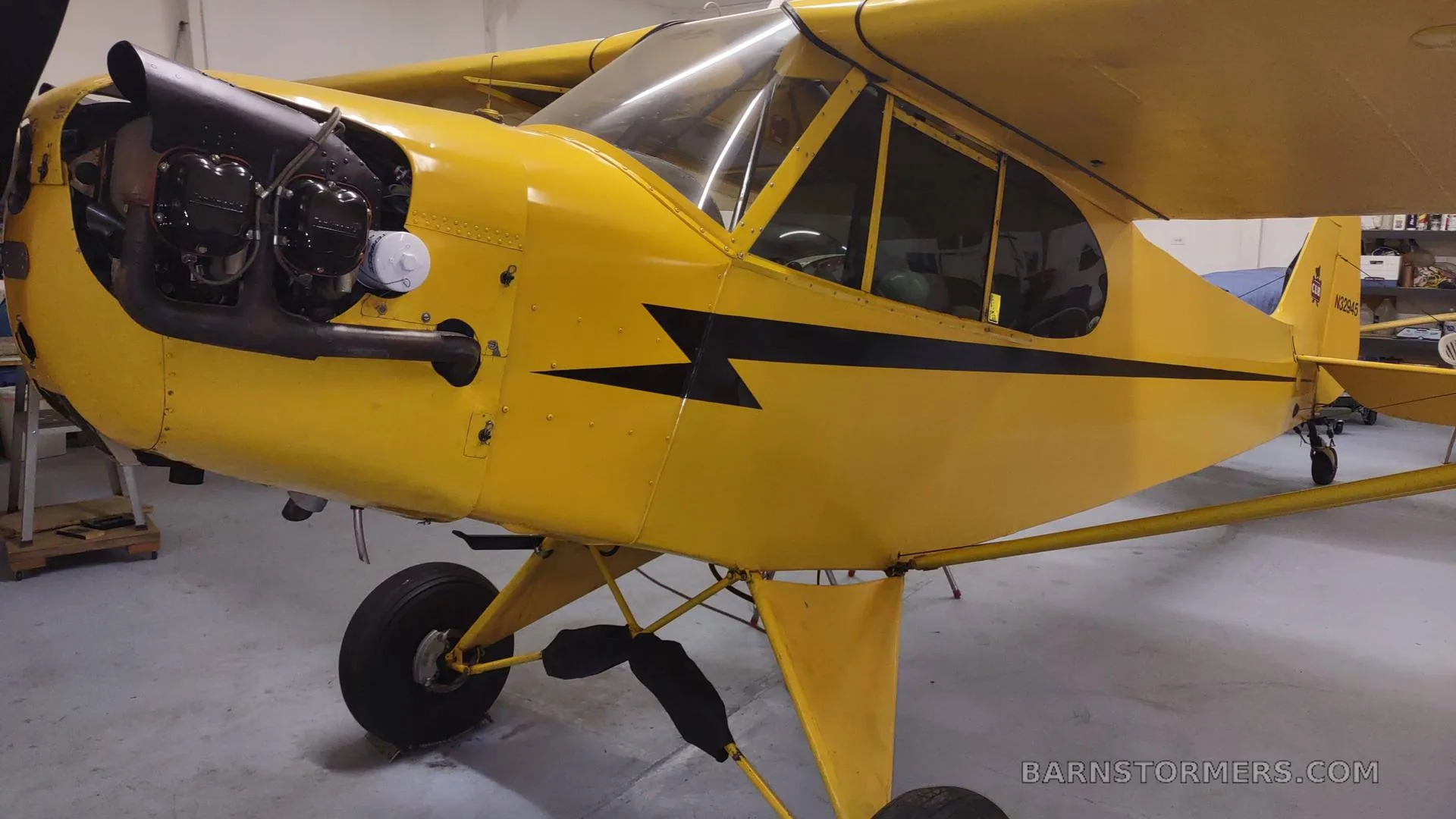 A yellow Piper Cub aircraft with a black stripe and exposed engine is parked in a hangar, viewed from the front left.