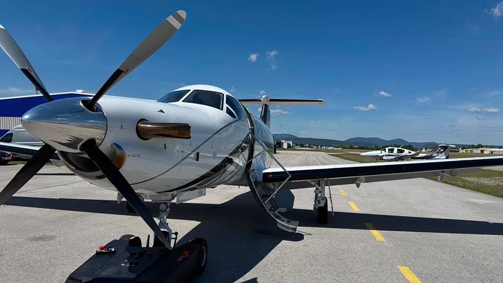 A white Pilatus PC-12 aircraft with its main cabin door open is parked on an asphalt tarmac with another aircraft in the background.