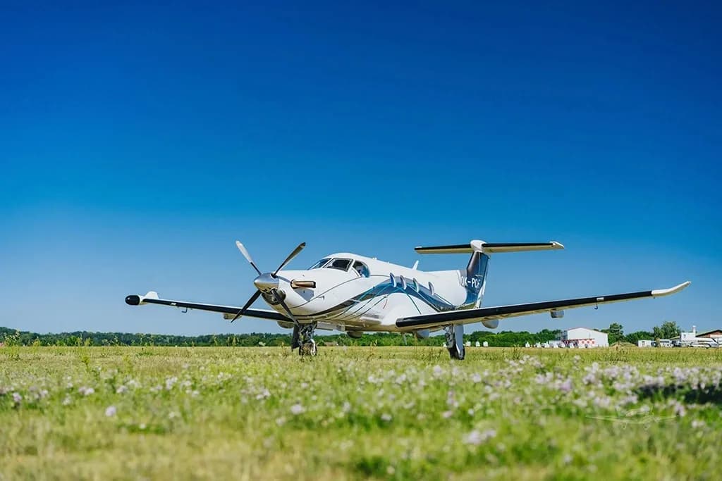 A white Pilatus PC-12 aircraft with blue and black accents is parked on a grassy field under a clear blue sky.
