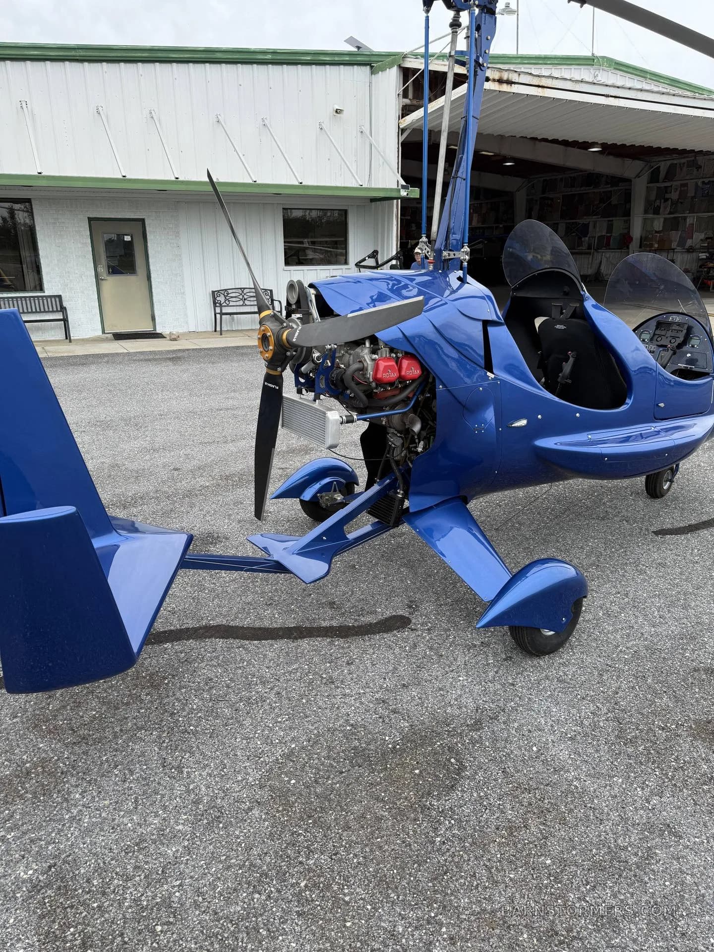 A blue autogyro is parked on an asphalt tarmac, viewed from the front-right, next to a white building.