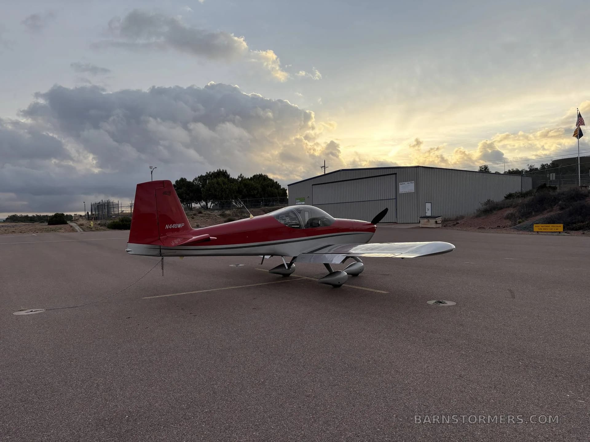 A red and silver single-engine aircraft, N448WP, tied down on an airfield with a hangar and sunset in the background.
