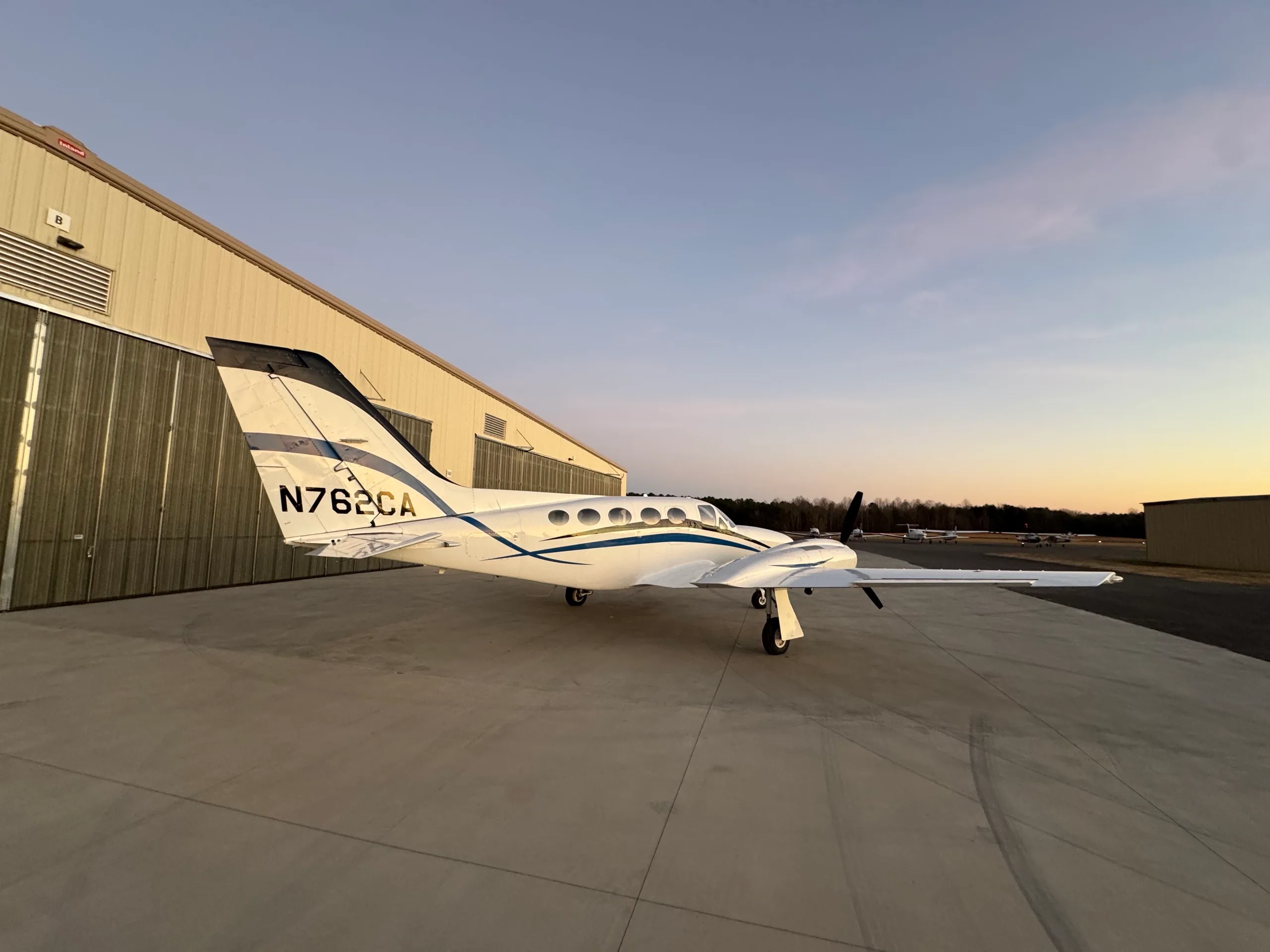 A white twin-engine aircraft with blue stripes, N762CA, parked beside a hangar at dusk.