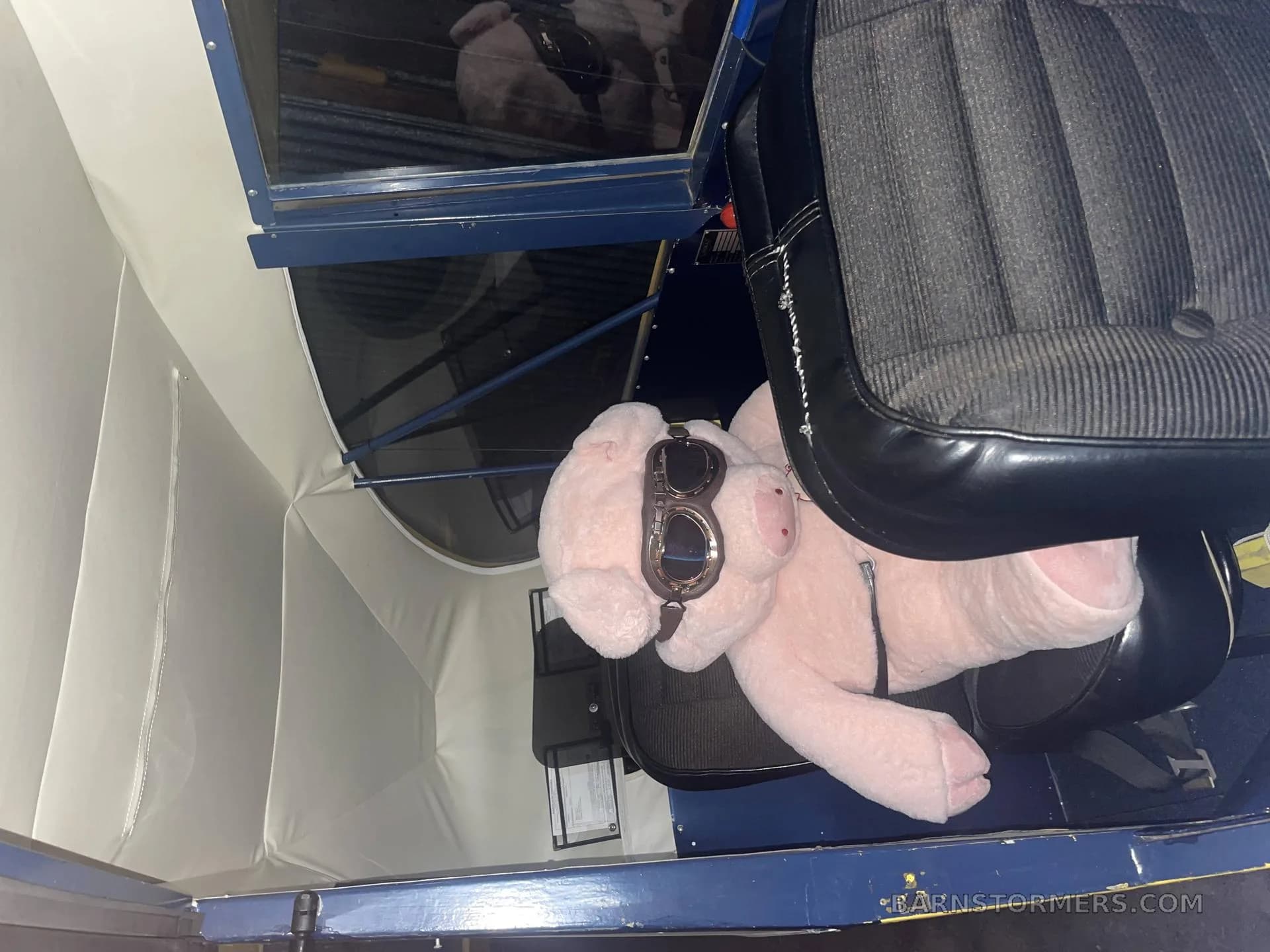 A pink pig plush toy wearing aviator goggles sitting in the passenger seat of an aircraft cabin.