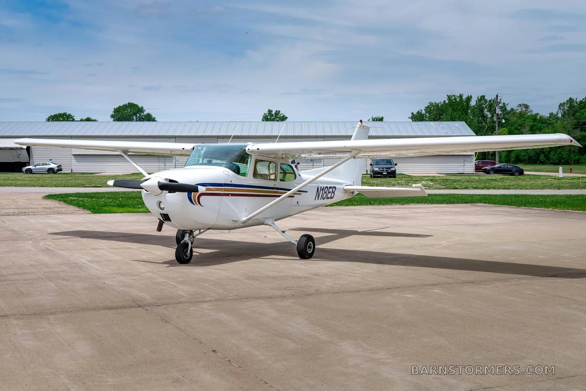 A white Cessna 172 (N18EB) with blue and gold stripes is parked on a tarmac, viewed from the front-left.