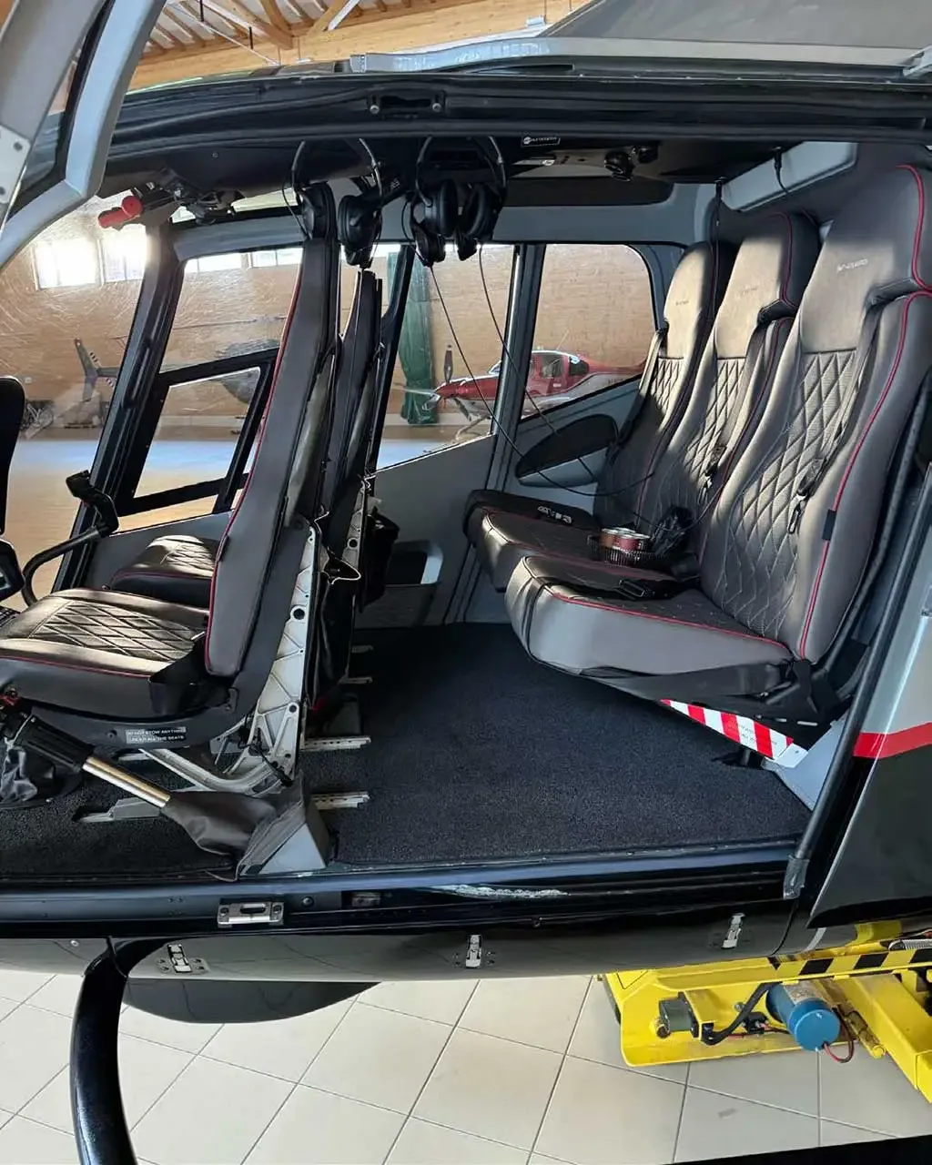 The interior cabin of a helicopter showing four grey quilted seats with red stitching and open doors.