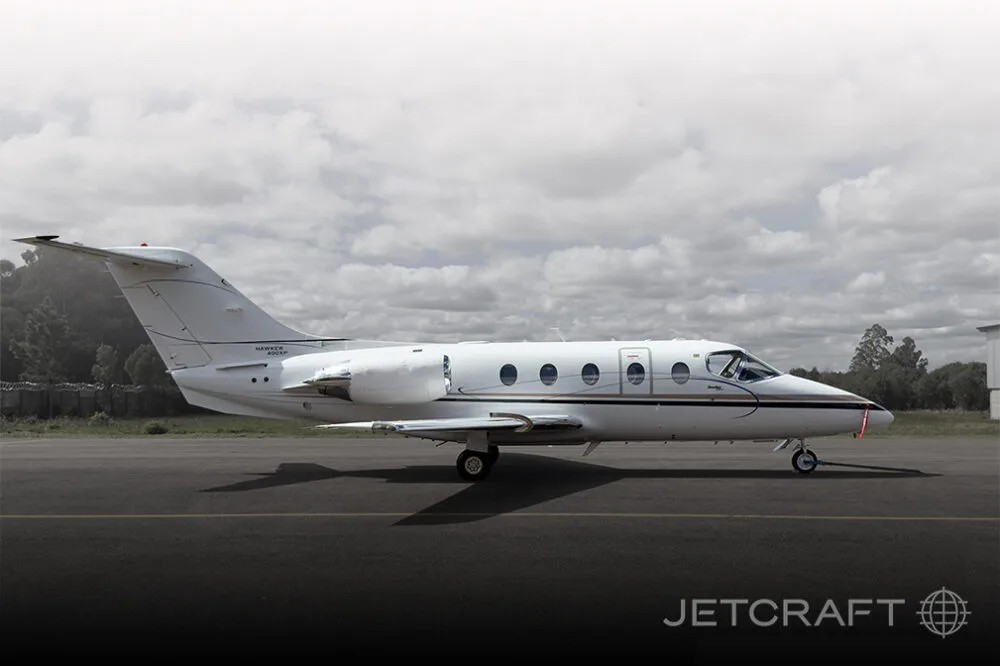 A white Hawker 800XP business jet with a dark stripe on the tarmac under a cloudy sky.