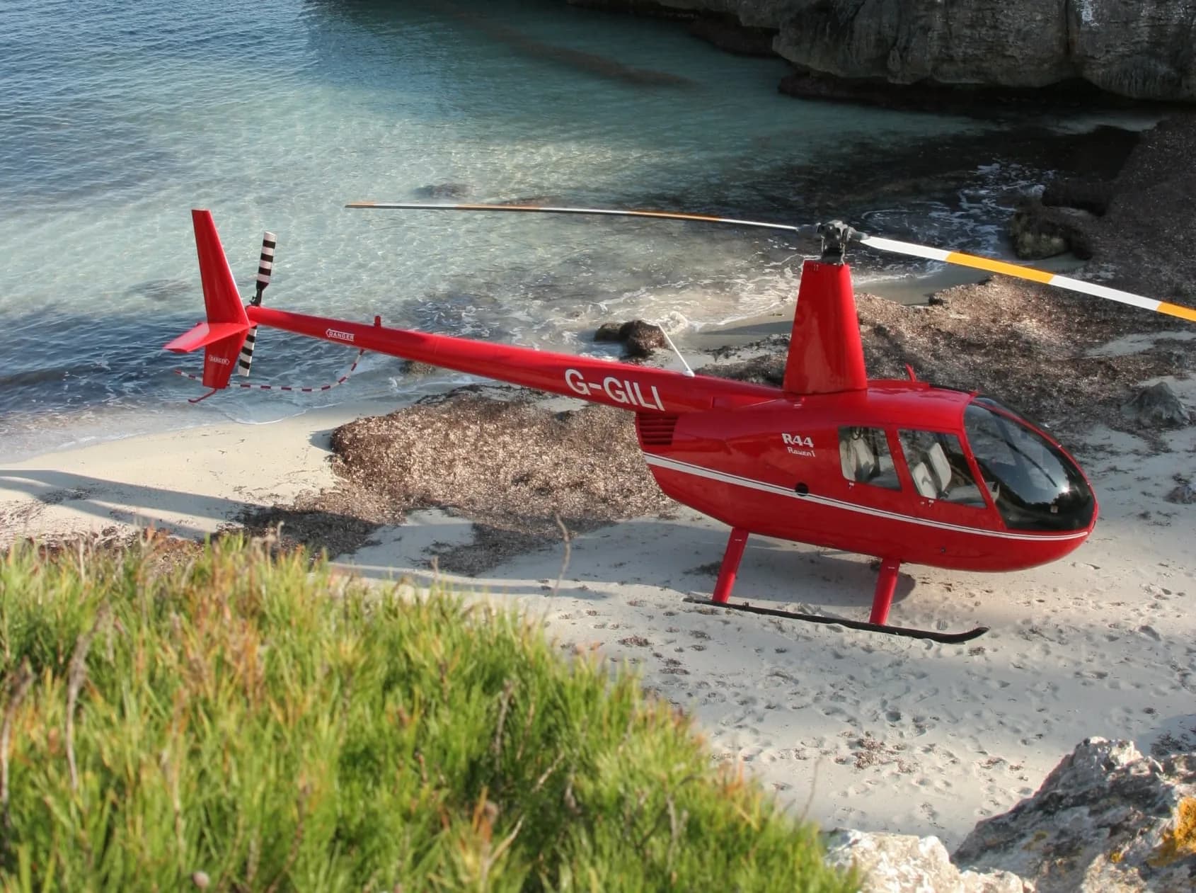 A red Robinson R44 helicopter parked on a beach with the registration G-GILI.