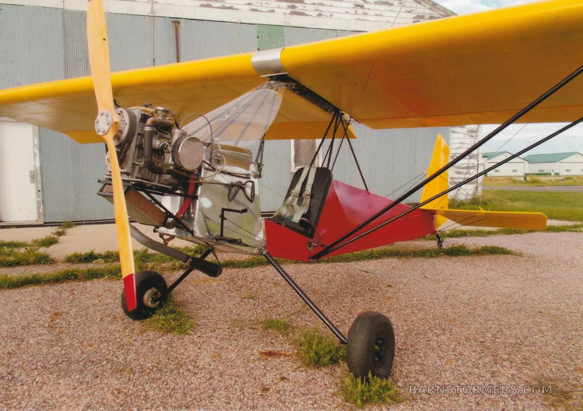 A side view of a yellow-winged, red-bodied ultralight aircraft with an exposed engine, parked on gravel in front of a hangar.