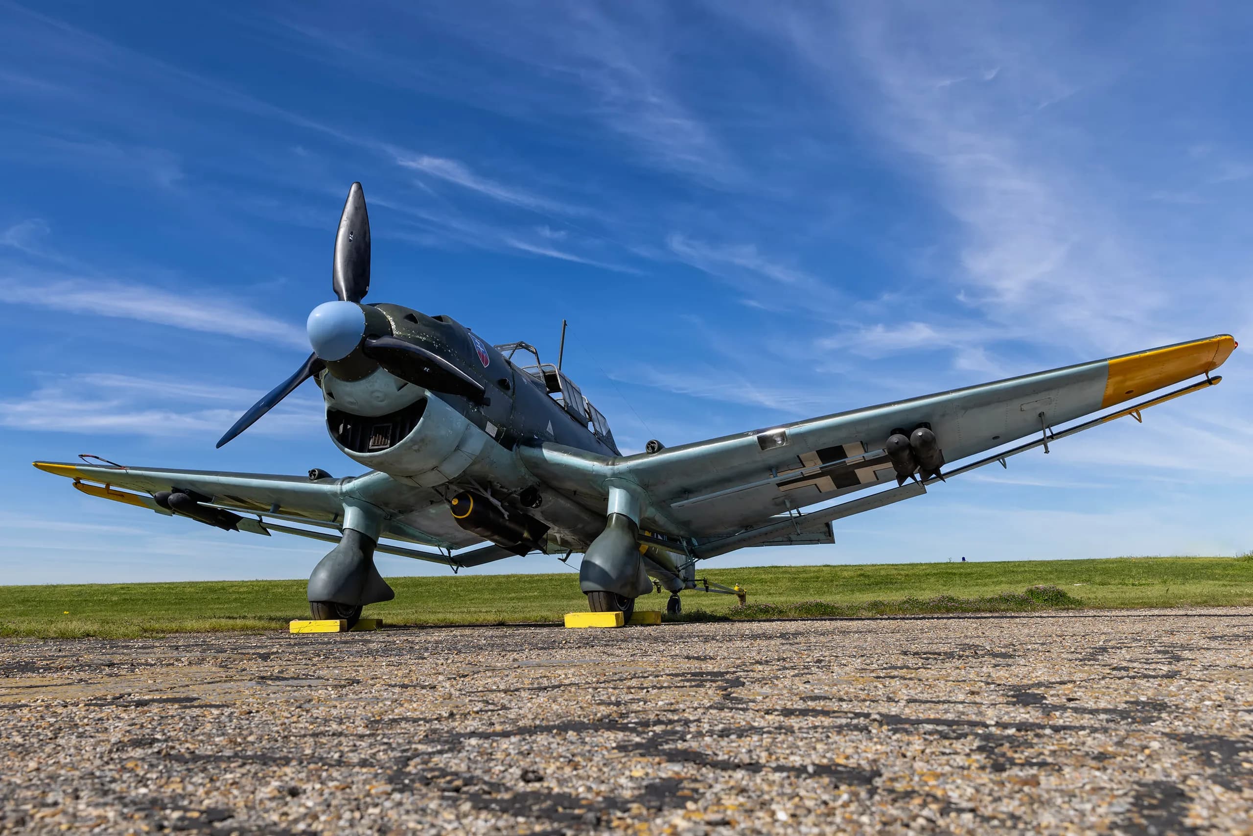 A low-angle view of a dark green and light blue Junkers Ju 87 Stuka dive bomber on an airfield under a blue sky.