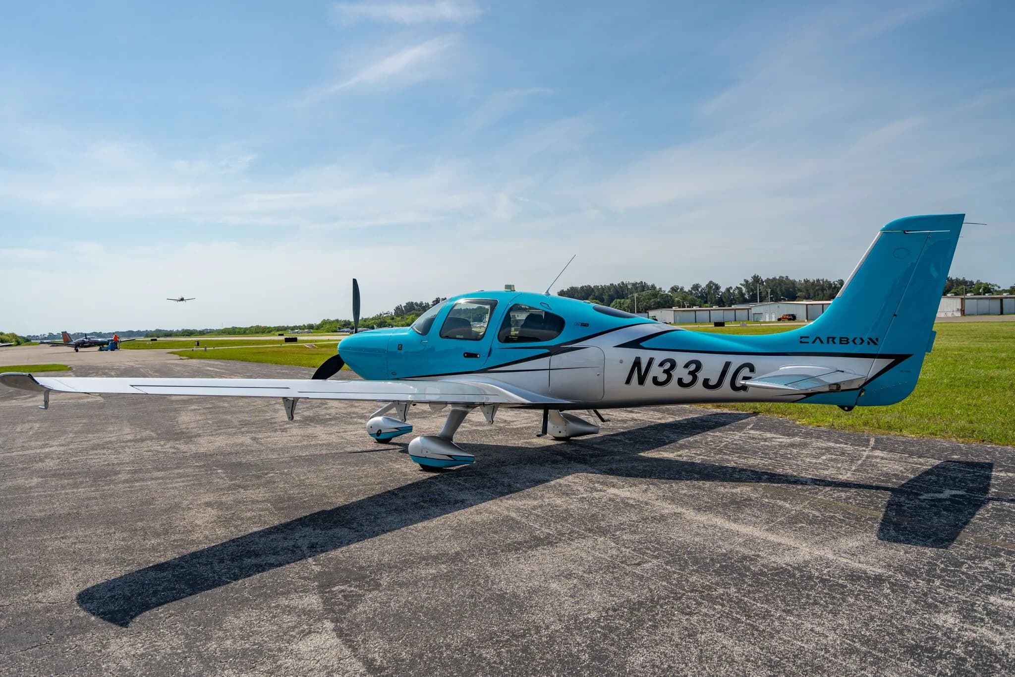 A blue and silver Carbon aircraft (N33JG) parked on an asphalt ramp with another aircraft in the distance.