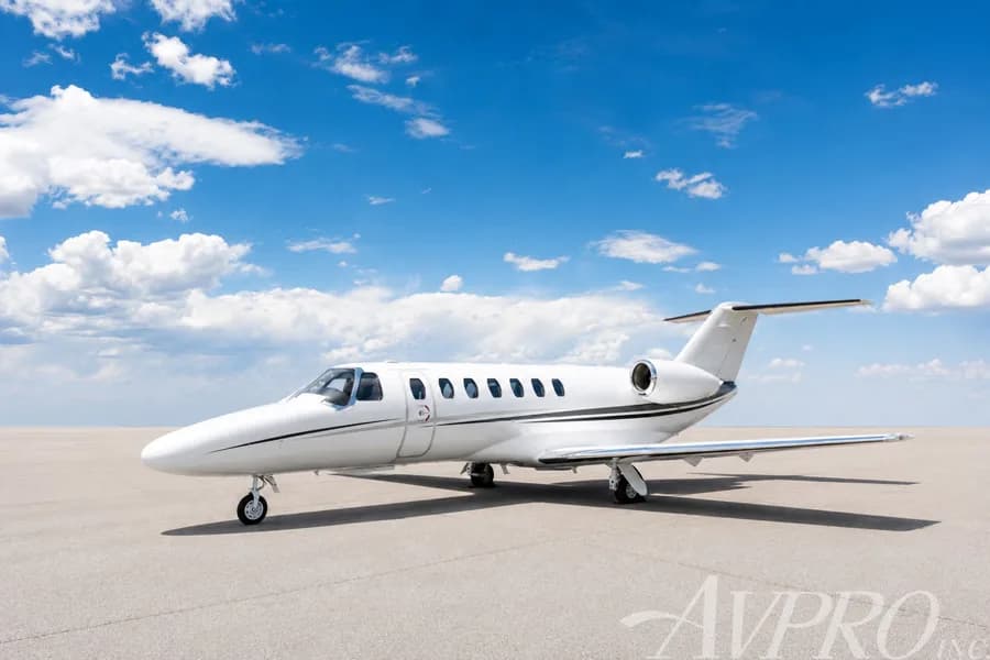 A white Cessna Citation jet with black and grey stripes parked on an airfield under a blue sky with clouds.