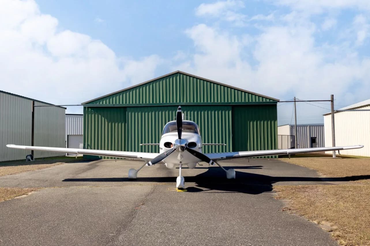 Front view of a white Cirrus SR22 aircraft with a three-blade propeller, parked in front of a green hangar.