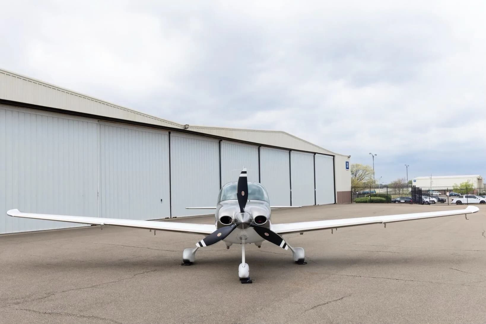A front view of a silver and white Cirrus SR22 aircraft with a three-blade propeller, parked on an tarmac.