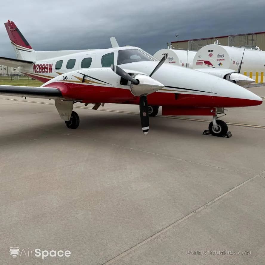 A white and red twin-engine propeller aircraft with gold accents, registration N2889W, parked on an asphalt ramp with fuel tanks in the background.