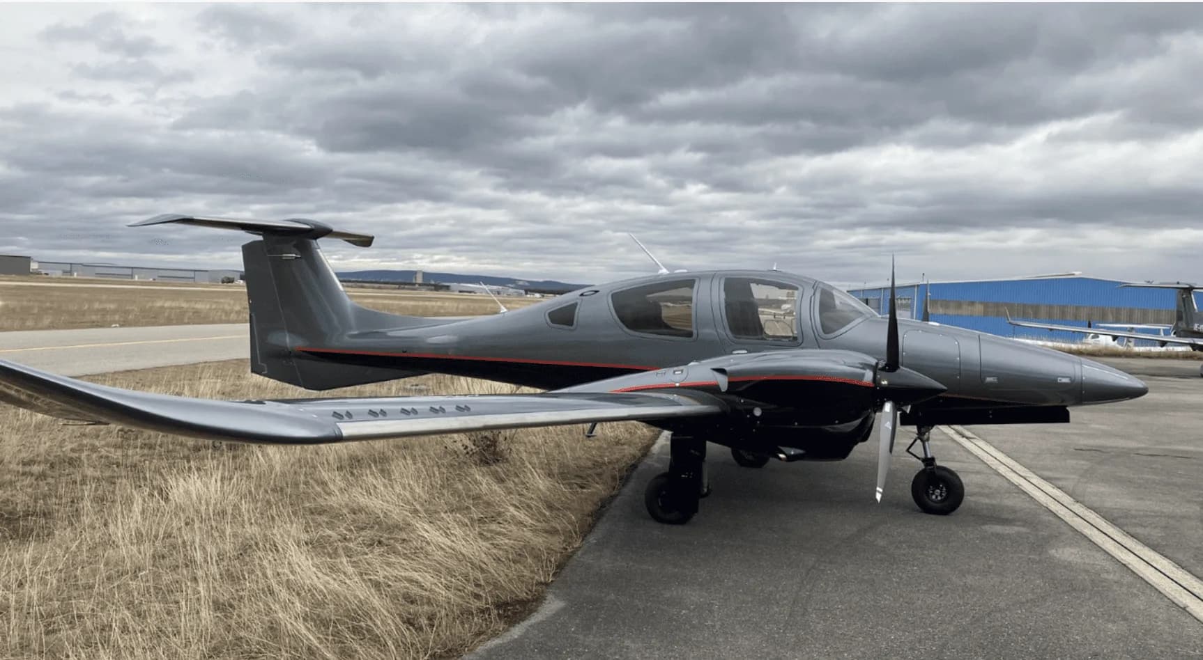A grey Diamond DA62 twin-engine aircraft with red pinstripes parked on a tarmac next to dry grass under a cloudy sky.