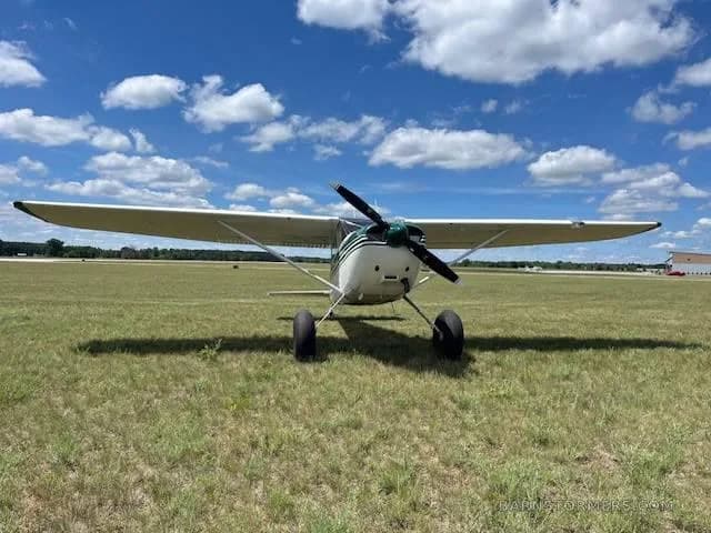 A front view of a white and green single-engine propeller aircraft with large tires, parked on a grassy field under a blue sky with clouds.