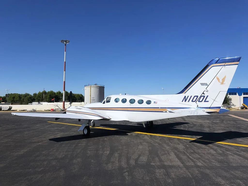 Rear-side view of a white Cessna 421 Golden Eagle aircraft with blue and gold stripes on the tarmac under a clear blue sky.