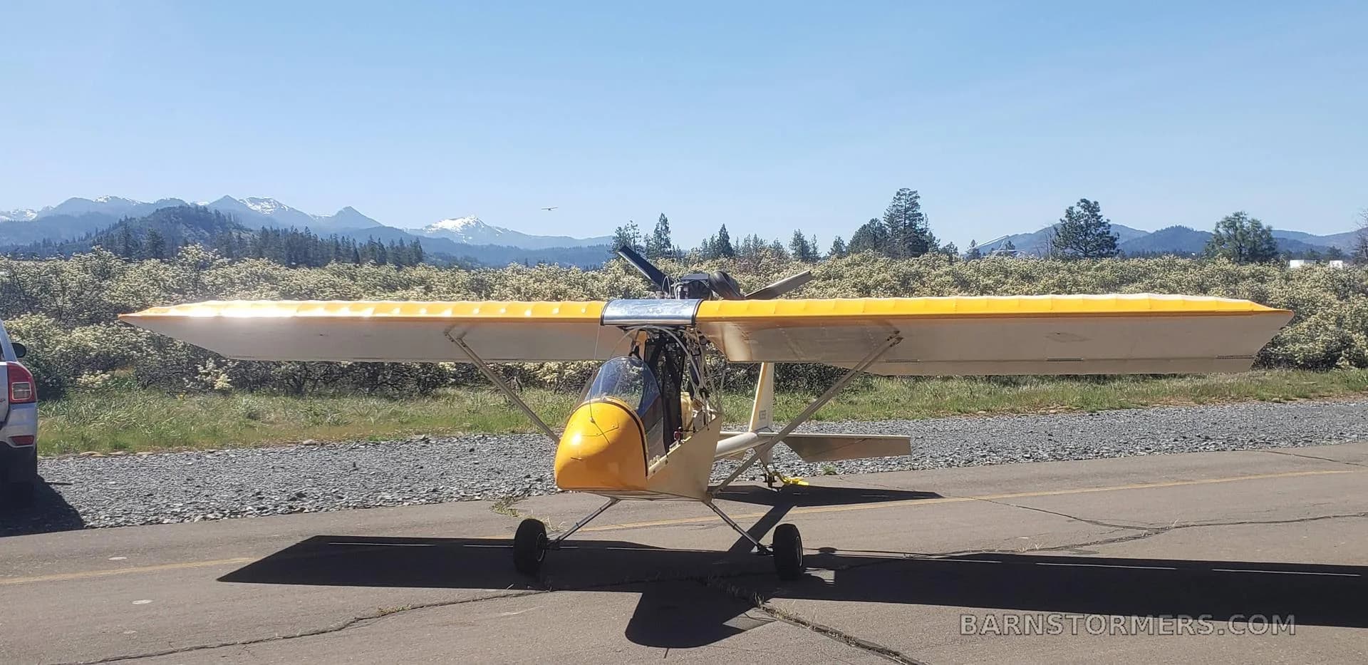 A yellow and white ultralight aircraft with a pusher propeller sits on an asphalt runway with mountains in the background.