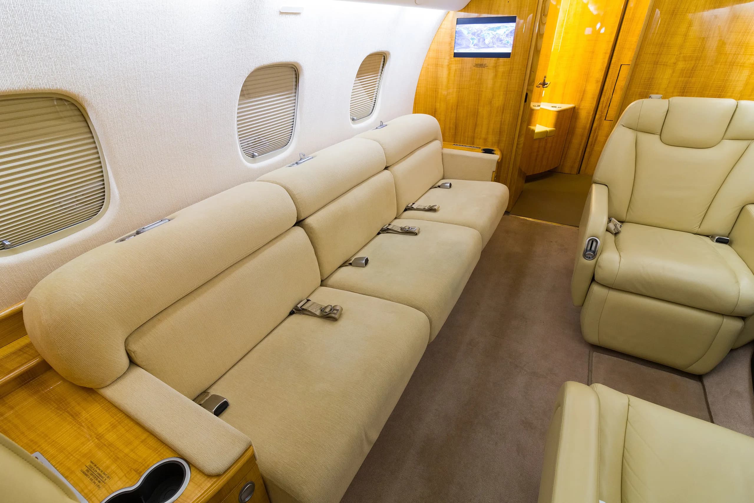 A private jet cabin interior featuring a long beige sofa and a single beige leather seat with polished wood accents.